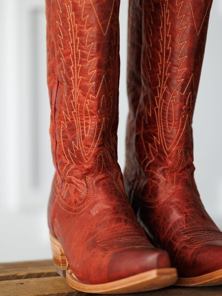 A close-up of The Pueblo tall, distressed leather western boots by Savannah Sevens western life{&}style, featuring decorative stitching and displayed on a wooden surface. Boots made by Black Star. 