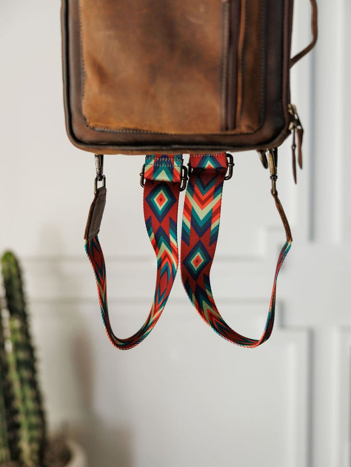 A close-up of The Ranch Hand backpack by Savannah Sevens western life{&}style, crafted in genuine leather with a red, teal, yellow, and black geometric strap. A potted cactus appears partially in the background.