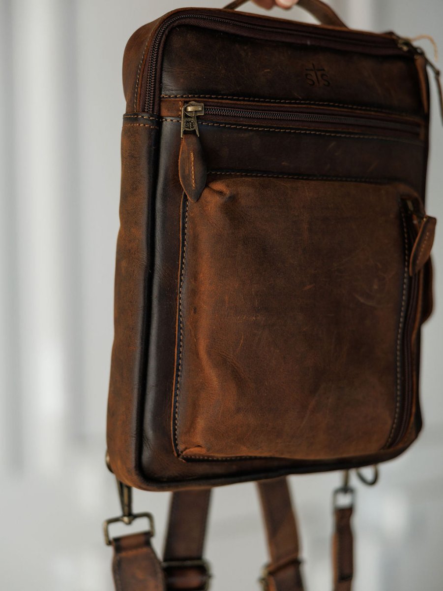 A close-up of The Ranch Hand by Savannah Sevens western life{&}style—genuine leather backpack with visible stitching, front zip pocket, and adjustable straps, designed as a laptop bag, hanging against a softly blurred background.