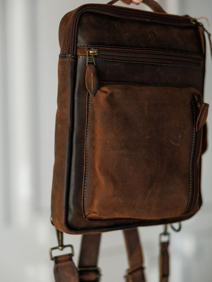 A close-up of The Ranch Hand by Savannah Sevens western life{&}style—genuine leather backpack with visible stitching, front zip pocket, and adjustable straps, designed as a laptop bag, hanging against a softly blurred background.