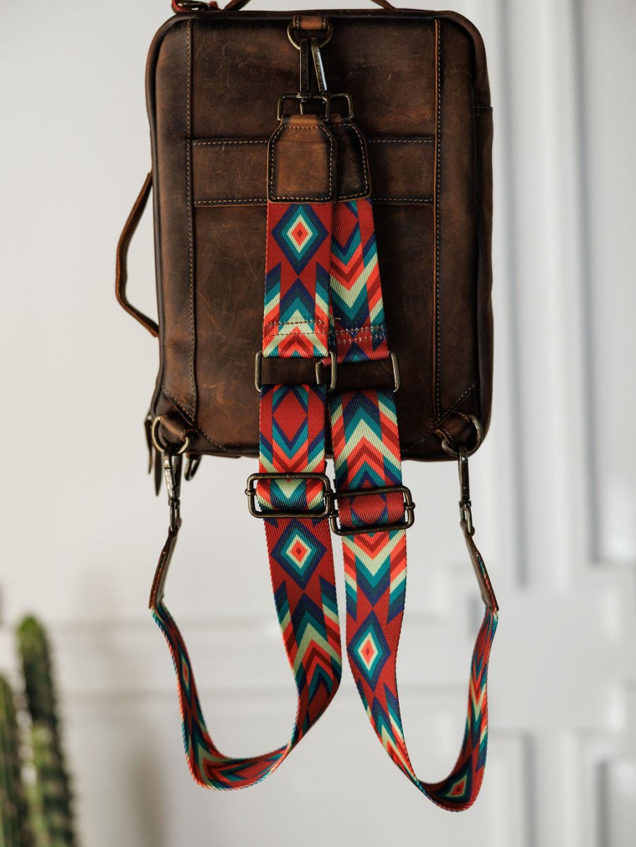 The Ranch Hand by Savannah Sevens western life{&}style, a brown leather laptop bag with a vibrant geometric strap in red, teal, blue, yellow, and white, hangs on a hook against a minimal white wall with a blurred plant in the background.