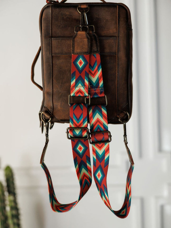 The Ranch Hand by Savannah Sevens western life{&}style, a brown leather laptop bag with a vibrant geometric strap in red, teal, blue, yellow, and white, hangs on a hook against a minimal white wall with a blurred plant in the background.