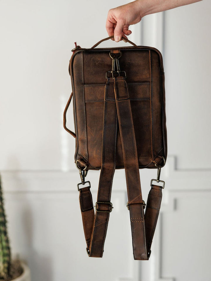 A hand holds up "The Ranch Hand" genuine leather backpack by Savannah Sevens western life{&}style. It features adjustable shoulder straps, brass hardware, and visible stitching. A blurred cactus appears in the lower left corner.