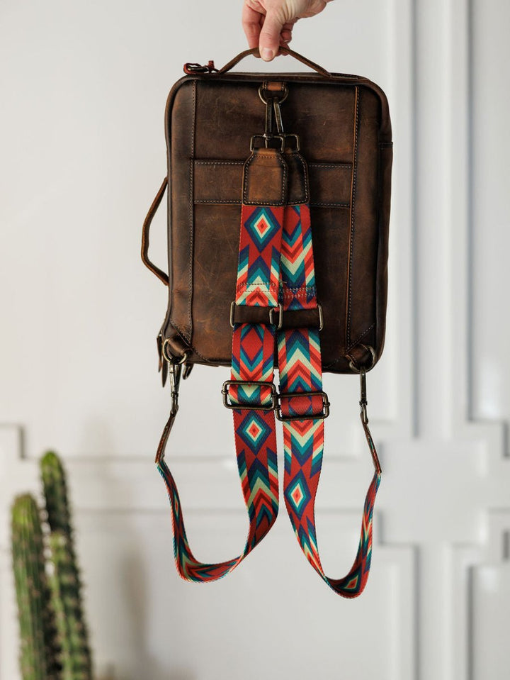 A hand holds up The Ranch Hand backpack by Savannah Sevens western life{&}style, made of brown genuine leather with a red, teal, and white geometric-patterned strap. A cactus sits in the lower left corner against a white panel wall background.