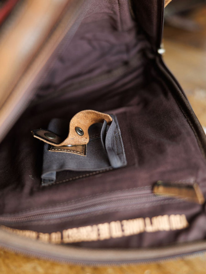 The Ranch Hand by Savannah Sevens western life{&}style displays its brown exterior, dark fabric lining, small black pocket, and leather buckle strap with metal snaps. A zipper and partial inscription are visible at the base of this genuine leather backpack.