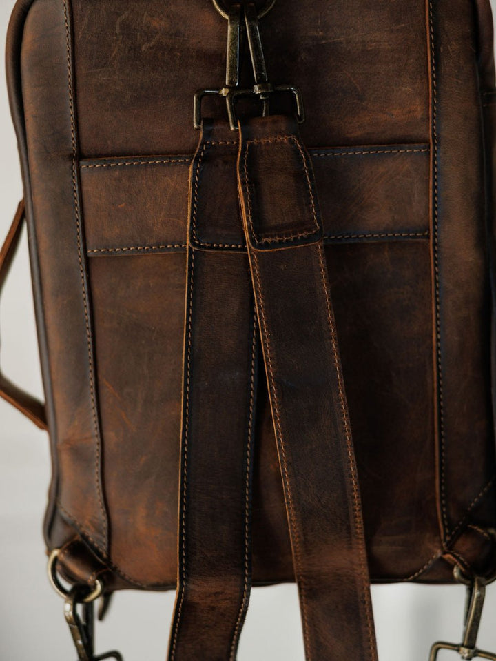 Close-up of The Ranch Hand backpack by Savannah Sevens western life{&}style, featuring genuine leather with a vintage look, stitched seams, adjustable shoulder straps, metal hardware, and a rectangular back pocket.
