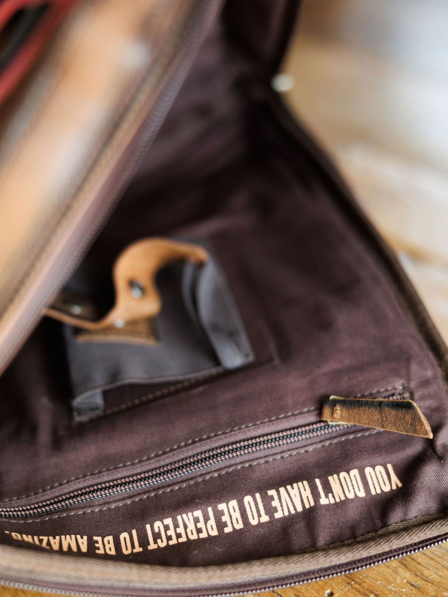 The Ranch Hand by Savannah Sevens western life{&}style is a brown leather laptop bag featuring a concealed carry holster, with “YOU DON’T HAVE TO BE PERFECT TO BE AMAZING” printed on the fabric lining near a zippered pocket.