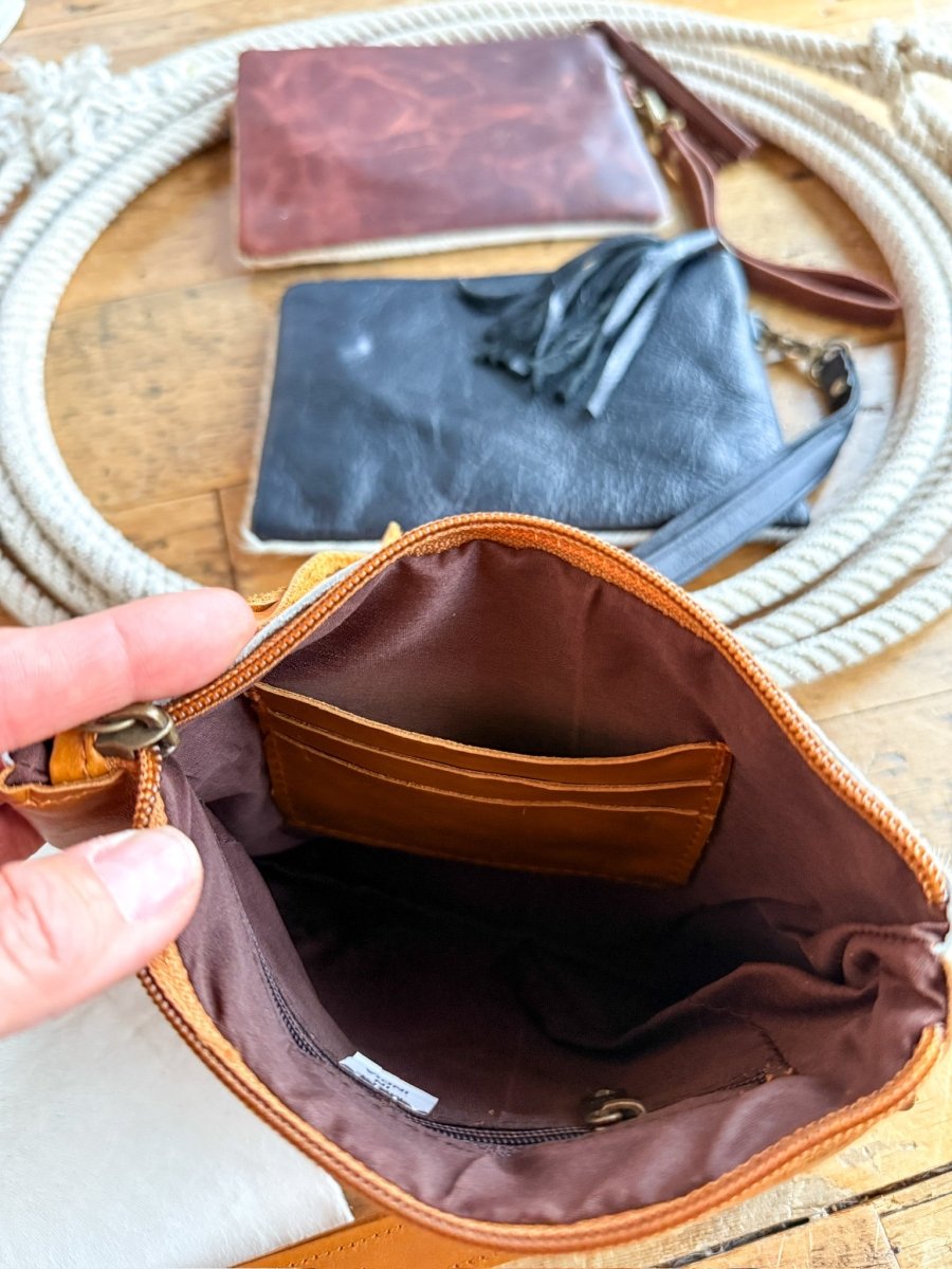 A hand displays the inner compartments of The Roanoke by Savannah Sevens western life{&}style, a small brown distressed leather purse. Behind it, brown and black cowhide clutch pouches rest on wood surrounded by coiled rope.