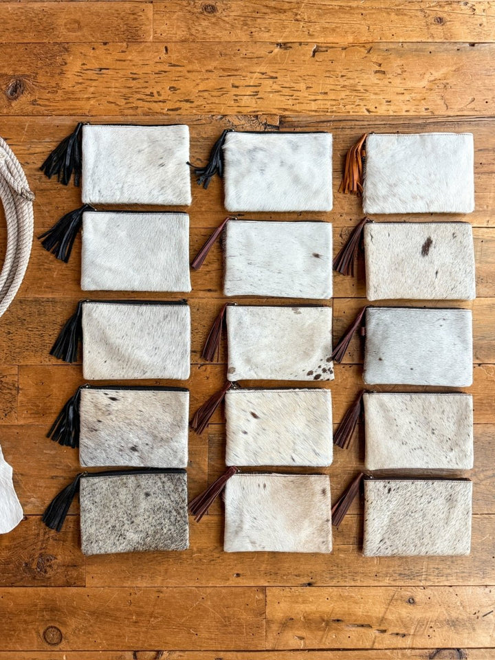 Fifteen "The Roanoke" cowhide clutch wallets by Savannah Sevens western life{&}style, each with a zipper and tassel, are arranged in a grid on wood. Every leather pouch displays unique natural markings.