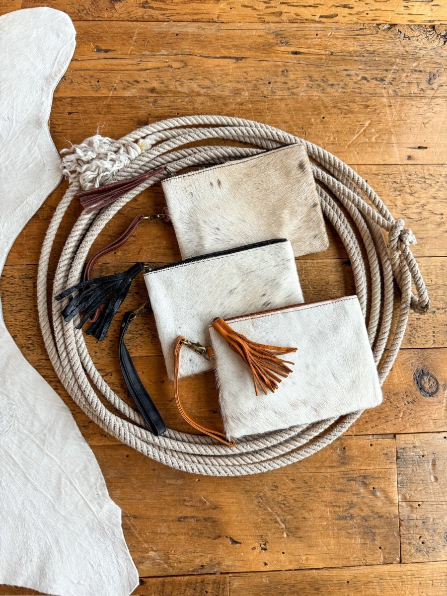 Three small Roanoke cowhide clutches with tassels by Savannah Sevens western life{&}style rest on a coiled rope atop rustic wood; a white fabric appears to the side. Each clutch’s distressed leather ensures unique character.