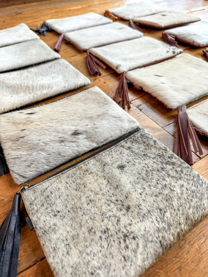Several rectangular pouches, called The Roanoke by Savannah Sevens western life{&}style, made from light-colored speckled cowhide with zipper closures and leather tassels are neatly arranged on a wooden floor, each clutch highlighting unique cowhide charm.