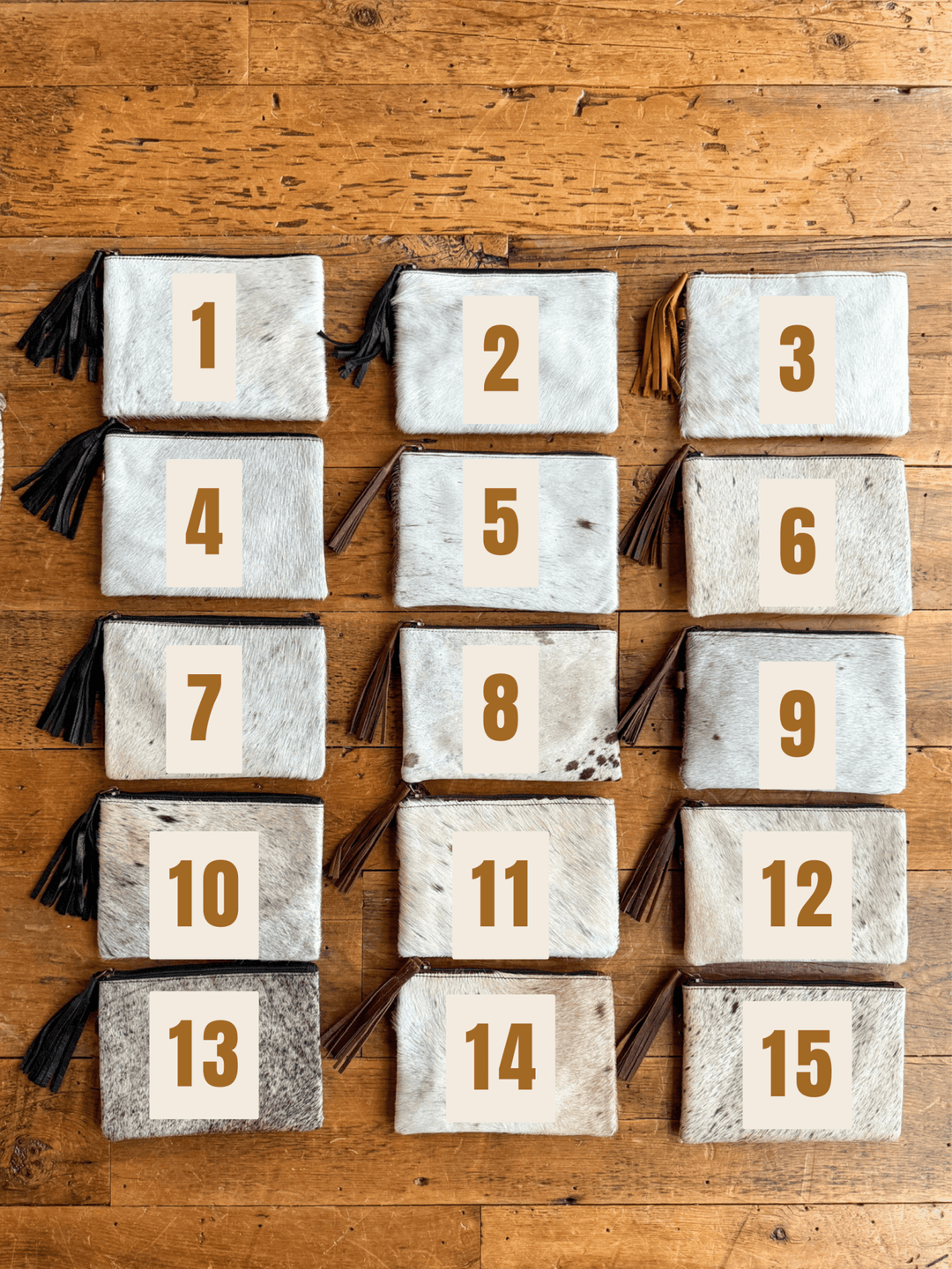 Fifteen "The Roanoke" cowhide clutch pouches by Savannah Sevens western life{&}style, each numbered 1–15 in large brown digits, are arranged in three columns on a wooden surface.