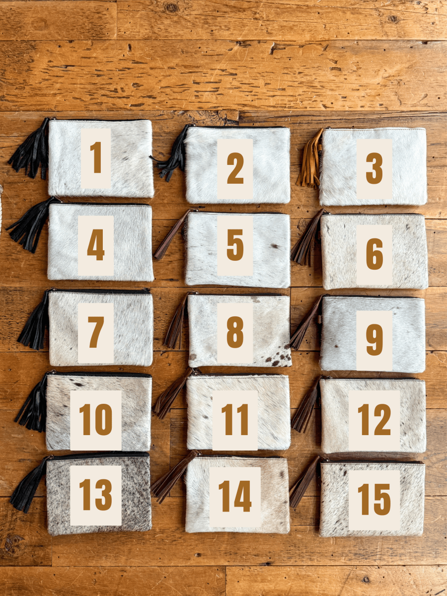Fifteen "The Roanoke" cowhide clutch pouches by Savannah Sevens western life{&}style, each numbered 1–15 in large brown digits, are arranged in three columns on a wooden surface.