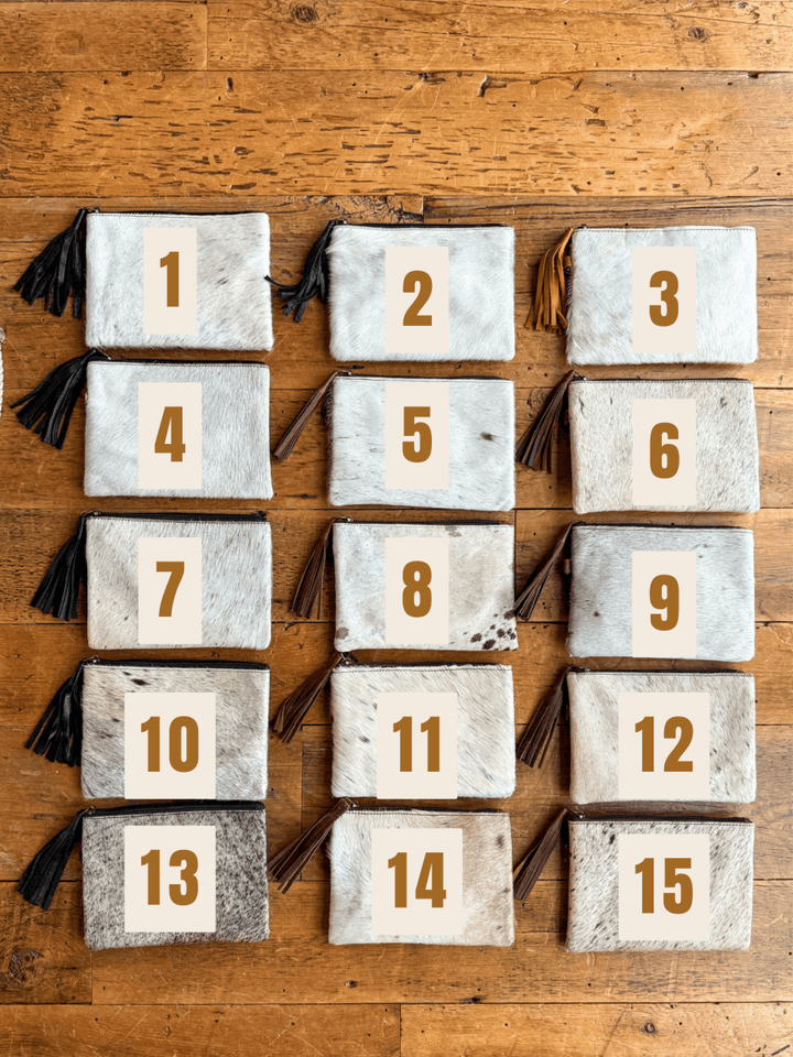 Fifteen "The Roanoke" cowhide clutch pouches by Savannah Sevens western life{&}style, each numbered 1–15 in large brown digits, are arranged in three columns on a wooden surface.
