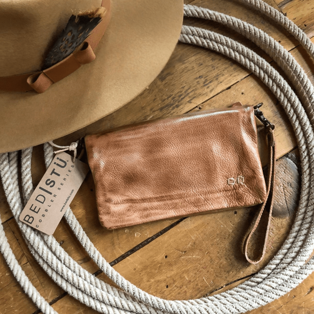 The Roca by Savannah Sevens western life{&}style, a tan dip-dye leather crossbody wristlet, sits on a wooden floor next to a coiled rope and a beige hat with a BEDSTU tag. By BedStu.