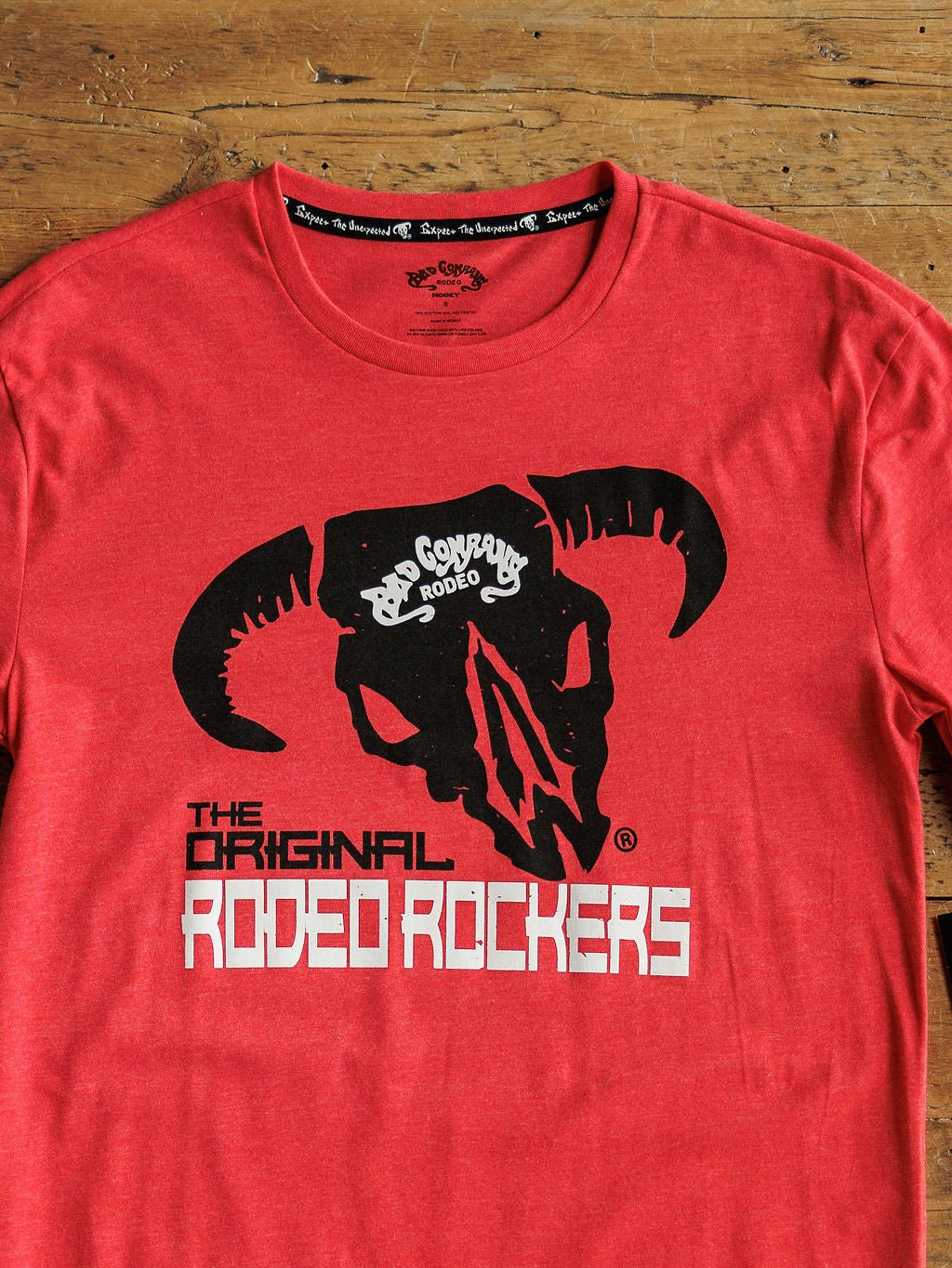 The Rodeo Rockers Men's Graphic Tee by Savannah Sevens western life{&}style features a bold black steer skull logo with "Bad Company Rodeo" and "Rip Cayway" above, and "The Original Rodeo Rockers" below, displayed on a red tee atop a wooden surface.
