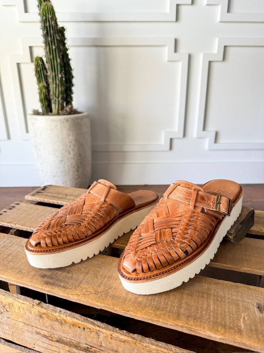 The Roswell by Savannah Sevens western life{&}style, woven leather huarache platform slip-ons with thick white soles, sits on a wooden pallet with a tall potted cactus and a white paneled wall in the background. Sandals by BedStu.
