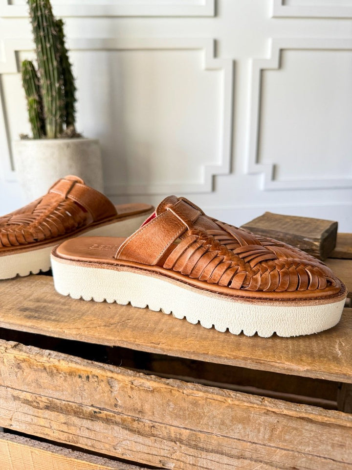 A pair of The Roswell tan woven leather platform slip-on sandals by Savannah Sevens western life{&}style, featuring thick white soles, displayed on rustic wood with a potted cactus and a white paneled wall in the background.Sandals made by BedStu. 