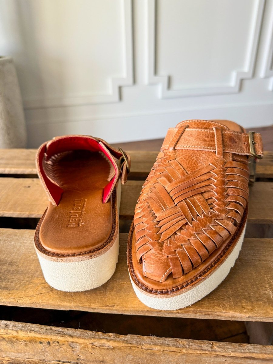 A pair of The Roswell woven leather huarache platform slip-on sandals by Savannah Sevens western life{&}style, with thick white soles—one showing the top, the other the open back and red interior—displayed on a wooden surface. Sandals made by BedStu. 