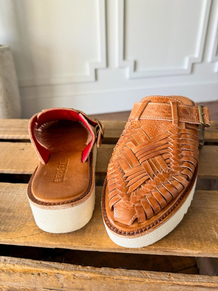 A pair of The Roswell woven leather huarache platform slip-on sandals by Savannah Sevens western life{&}style, with thick white soles—one showing the top, the other the open back and red interior—displayed on a wooden surface. Sandals made by BedStu. 