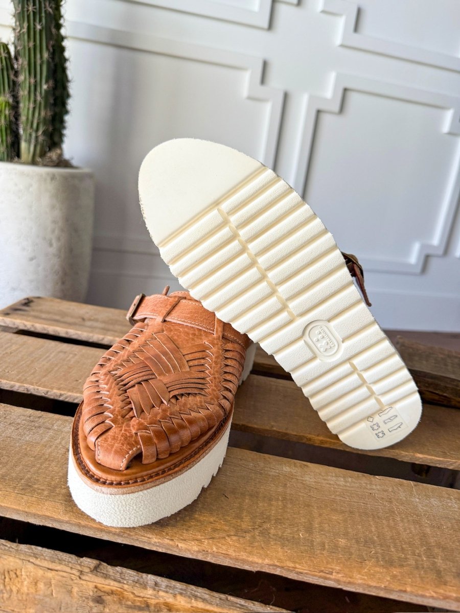 A pair of Savannah Sevens western life{&}style The Roswell woven leather huarache platform slip-on sandals with thick white rubber soles are shown on wood, one upright and one showing the textured sole. A potted cactus sits before a white wall in the background. Sandals made by BedStu. 