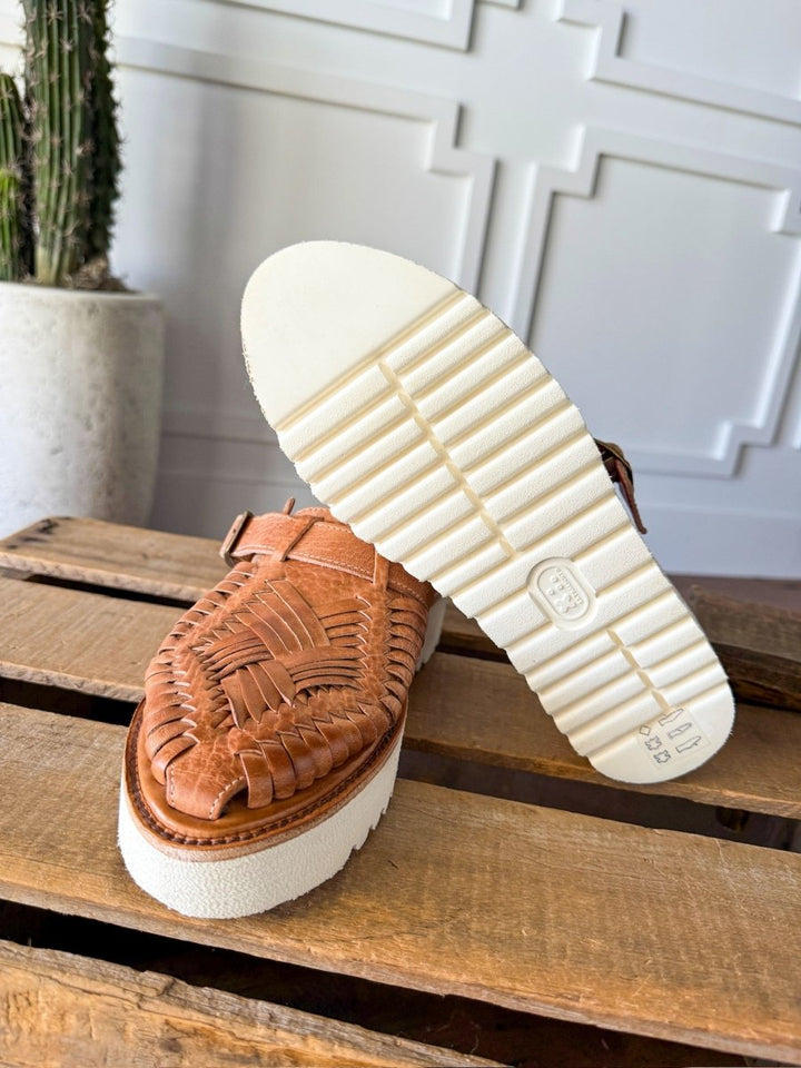 A pair of Savannah Sevens western life{&}style The Roswell woven leather huarache platform slip-on sandals with thick white rubber soles are shown on wood, one upright and one showing the textured sole. A potted cactus sits before a white wall in the background. Sandals made by BedStu. 