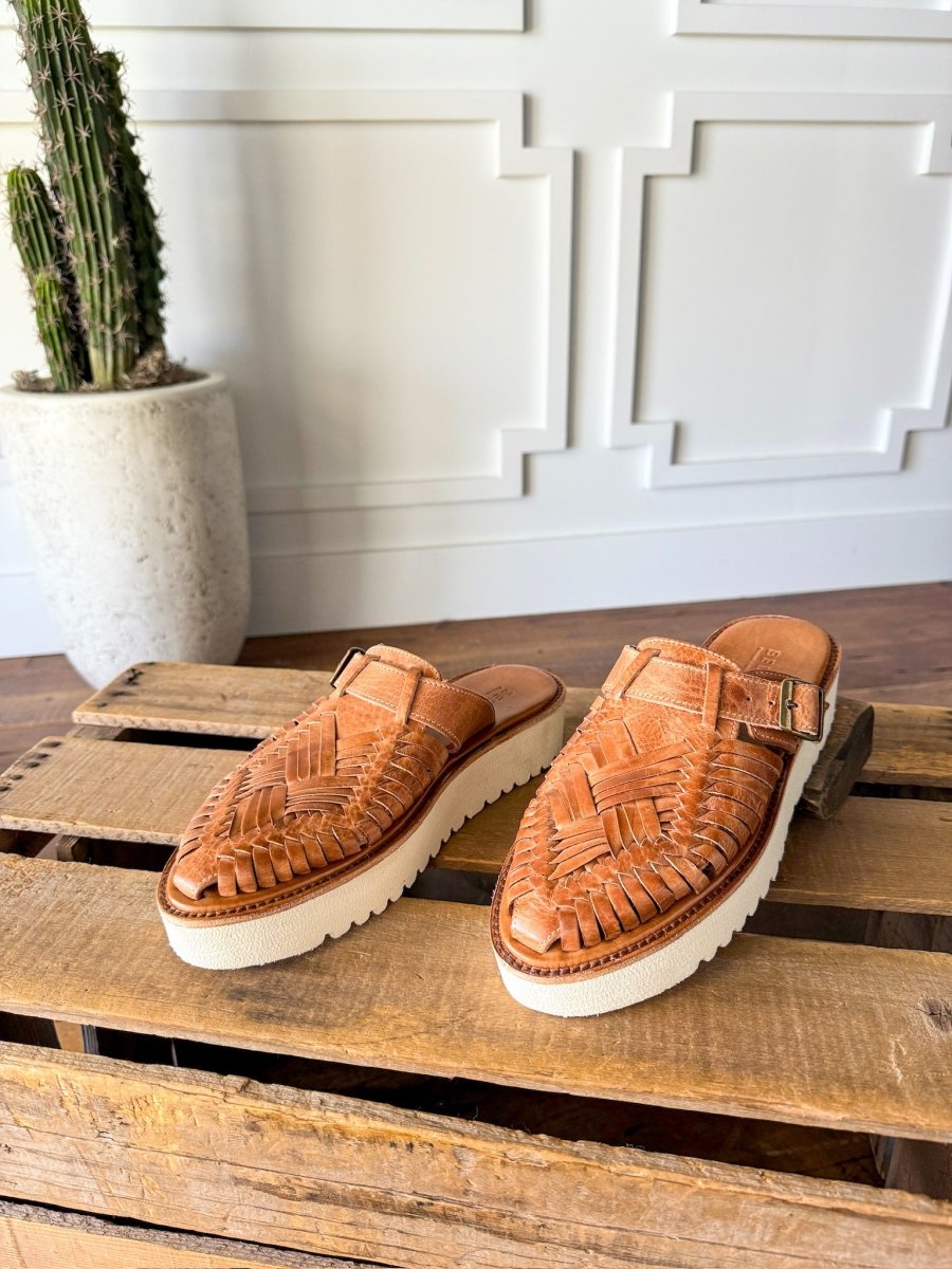 The Roswell slip-on woven leather huarache platforms by Savannah Sevens western life{&}style, featuring white soles, are displayed on a wooden crate with a tall potted cactus in the background against a white paneled wall. Sandals made by BedStu. 