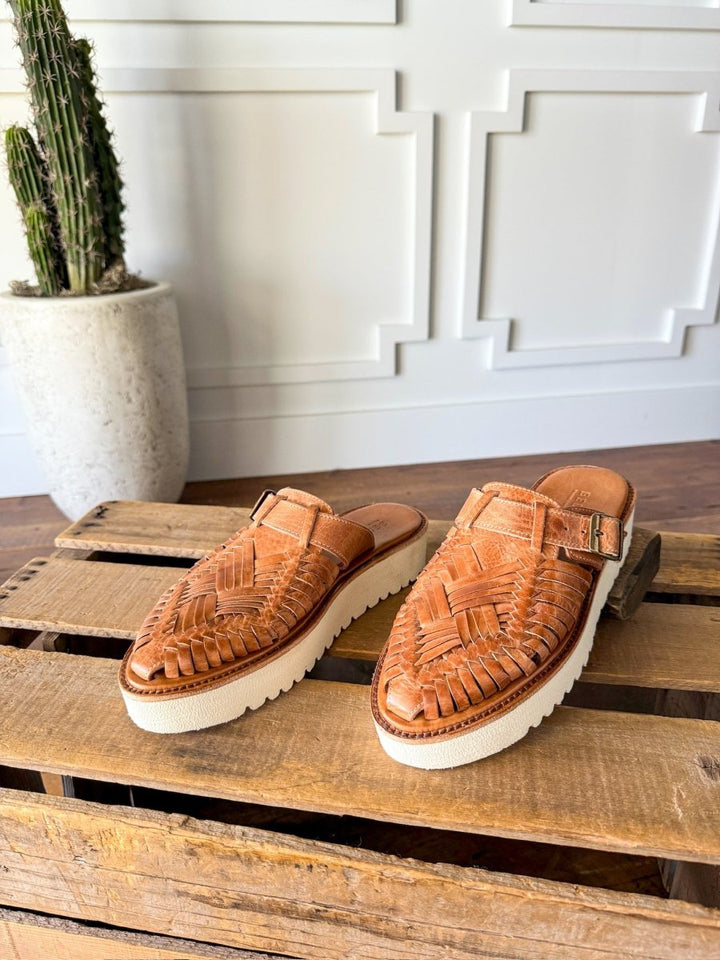 The Roswell slip-on woven leather huarache platforms by Savannah Sevens western life{&}style, featuring white soles, are displayed on a wooden crate with a tall potted cactus in the background against a white paneled wall. Sandals made by BedStu. 