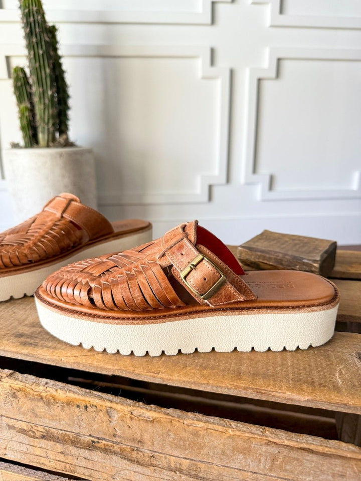 The Roswell by Savannah Sevens western life{&}style—a woven leather huarache platform slip-on sandal with a white sole and gold buckle—displayed on rustic wood with a potted cactus and a decorative white panel wall in the background. Sandals made by BedStu. 