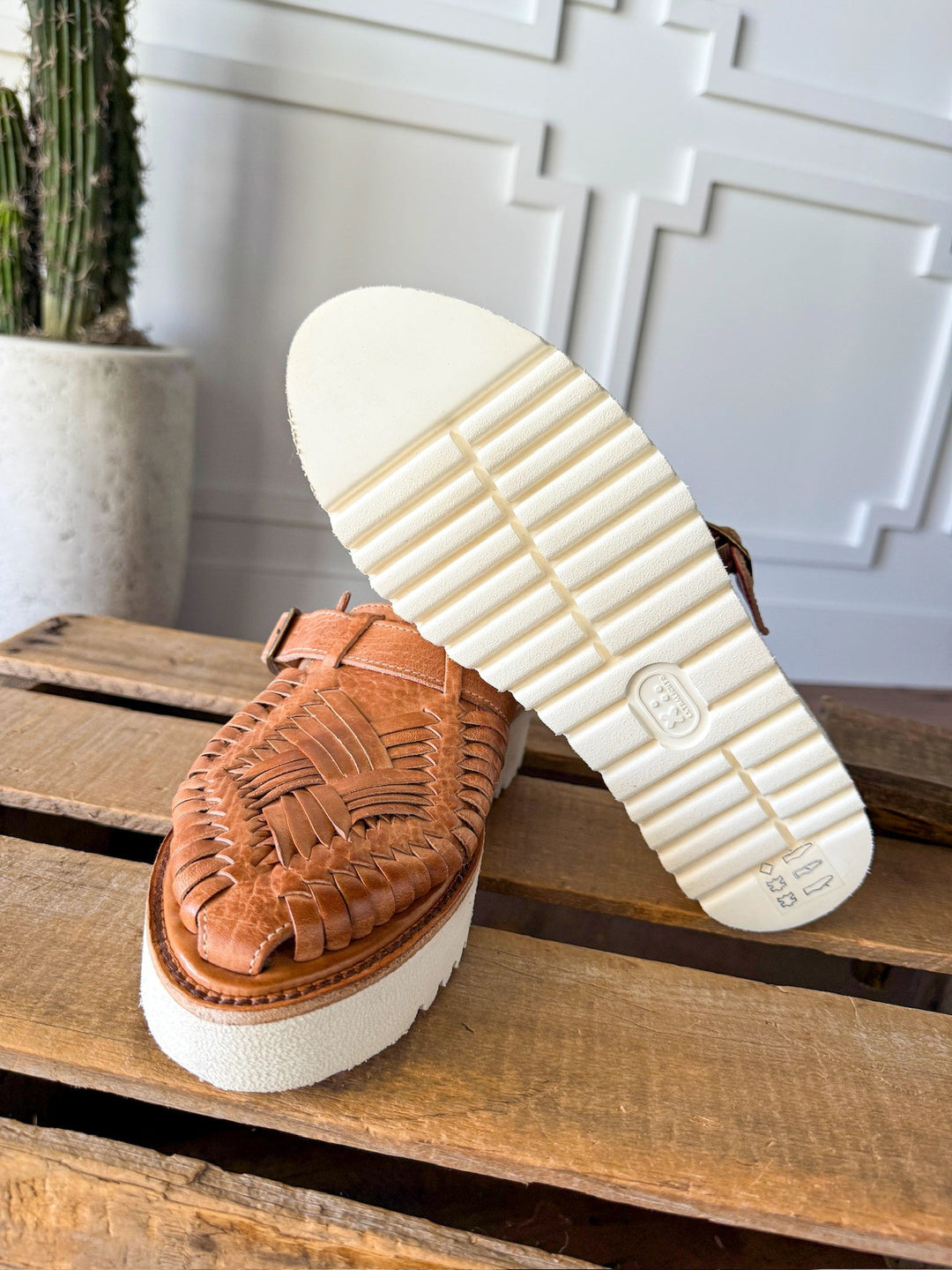 A pair of Savannah Sevens western life{&}style The Roswell woven leather huarache platform slip-on sandals with thick white rubber soles are shown on wood, one upright and one showing the textured sole. A potted cactus sits before a white wall in the background. Sandals made by BedStu. 