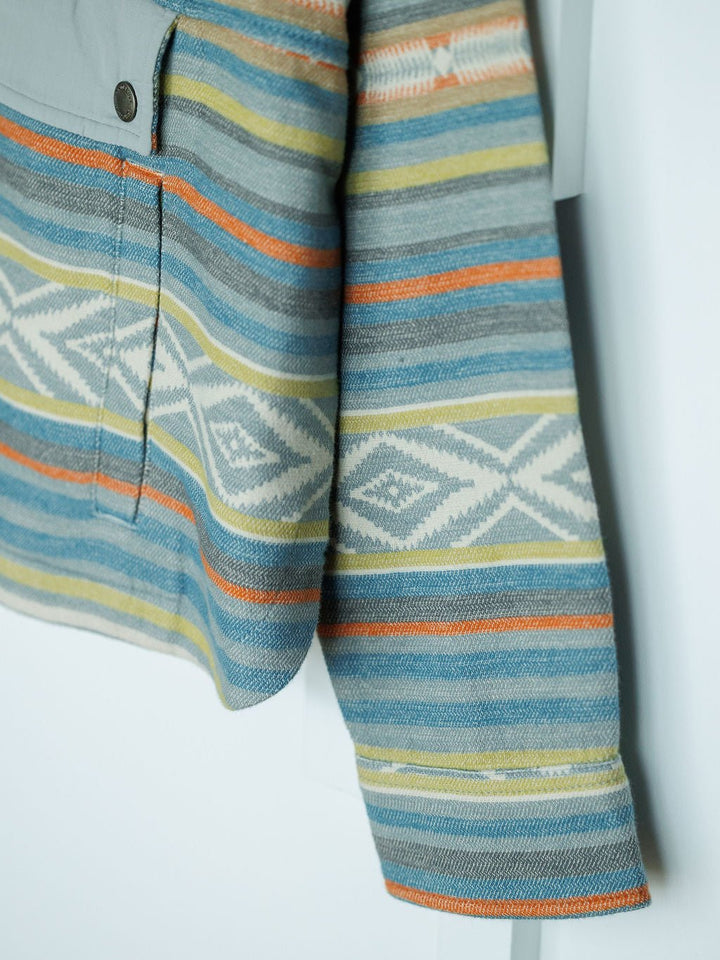A close-up of The Sallisaw by Savannah Sevens western life{&}style, a southwest quarter snap long-sleeve shirt with blue, orange, yellow, and white horizontal stripes and a geometric diamond pattern, hanging against a light background.