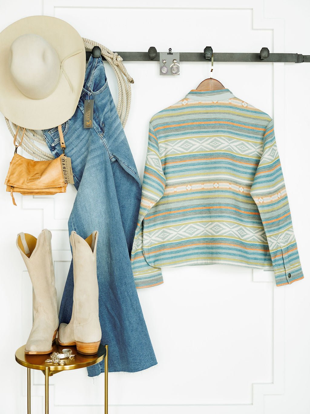 The Sallisaw by Savannah Sevens western life{&}style, featuring a beige hat, striped jacket, blue jeans, light tan cowboy boots, tan purse, and Pendleton pullover on a black wall rack with boots on a small gold table against a white background.