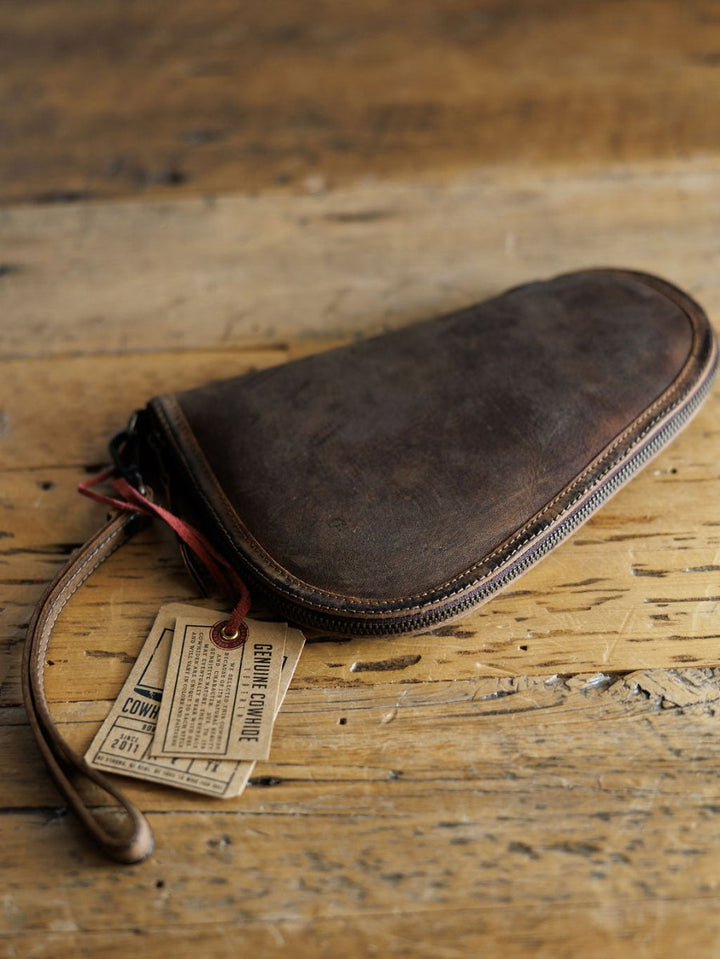The San Antonio by Savannah Sevens western life{&}style is a brown leather pistol case with a wrist strap, resting on rustic wood. Attached paper tags highlight its classic style.