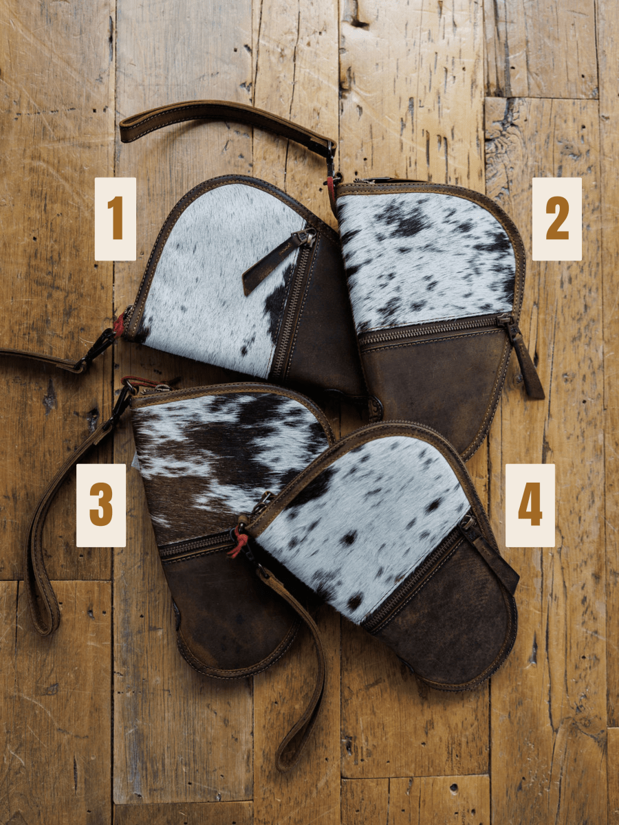 Four "The San Antonio" clutches from Savannah Sevens western life{&}style are displayed on a wooden floor, each labeled 1-4 and featuring distinctive white, brown, and black cowhide patterns.