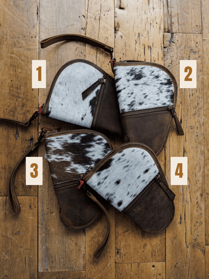 Four "The San Antonio" clutches from Savannah Sevens western life{&}style are displayed on a wooden floor, each labeled 1-4 and featuring distinctive white, brown, and black cowhide patterns.