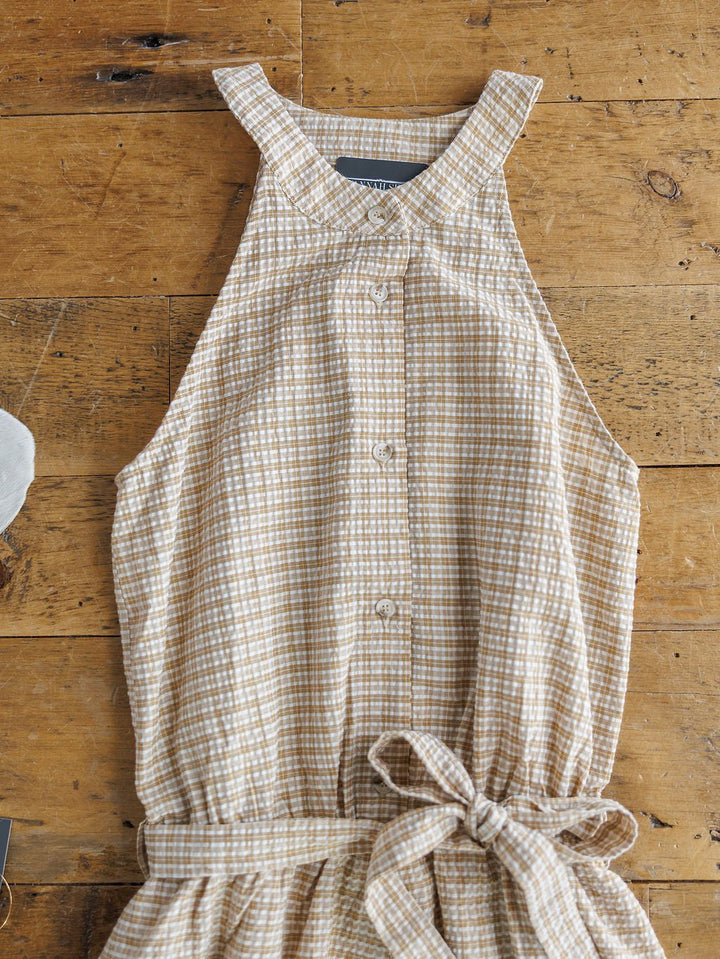 The Sanborn by Savannah Sevens western life{&}style is a sleeveless romper featuring a beige and white checkered pattern, high neckline, and tied waist, shown laid flat on a wooden surface.