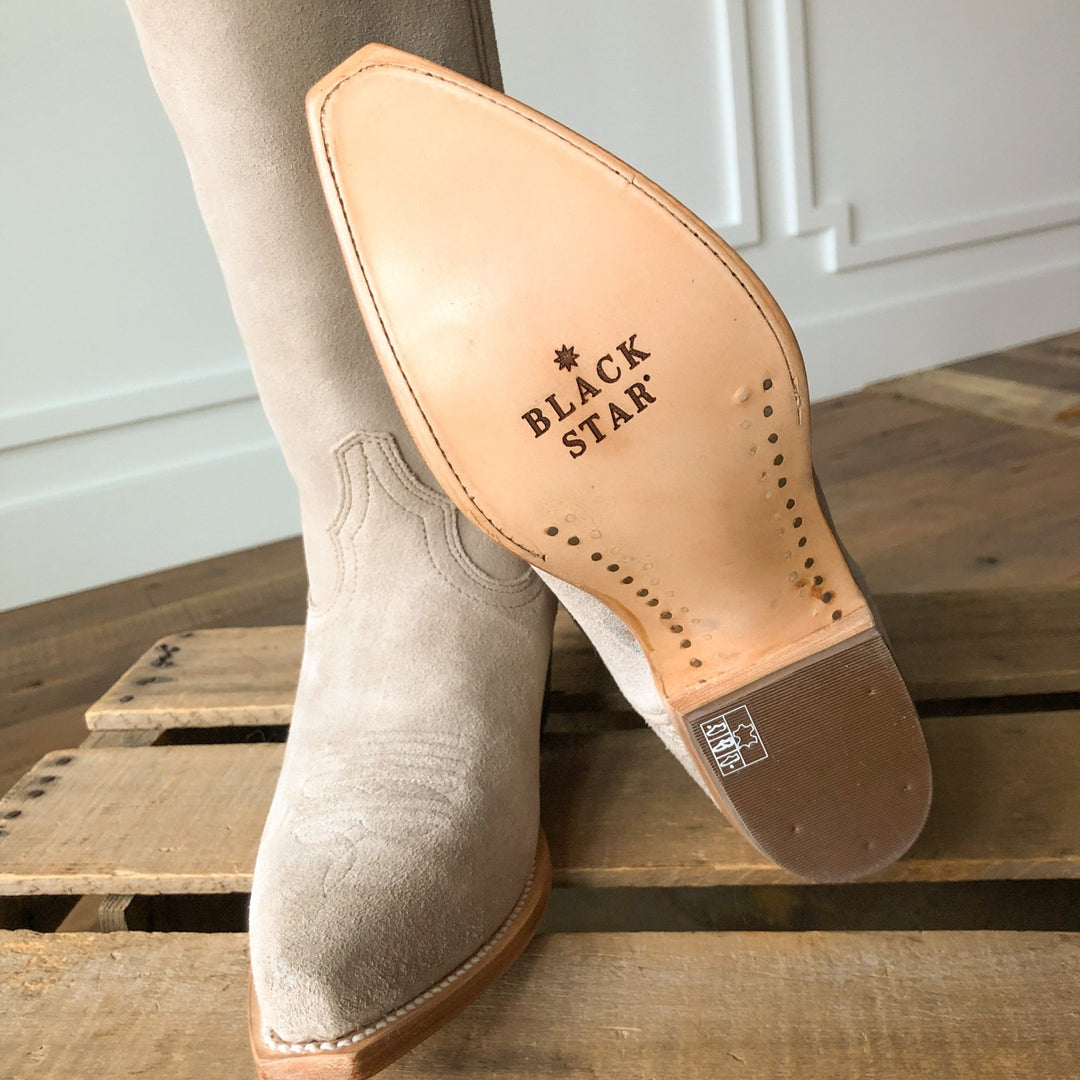 Close-up of The Shiloh, a light beige suede western boot with a snip toe from Savannah Sevens western life{&}style, resting on a wooden indoor surface. Made by Black Star. 