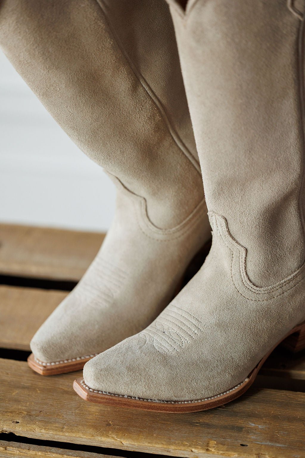 A close-up of The Shiloh western boots by Savannah Sevens western life{&}style, featuring light beige rough out leather, square toes, and decorative stitching, resting on a wooden surface. Made by Black Star. 
