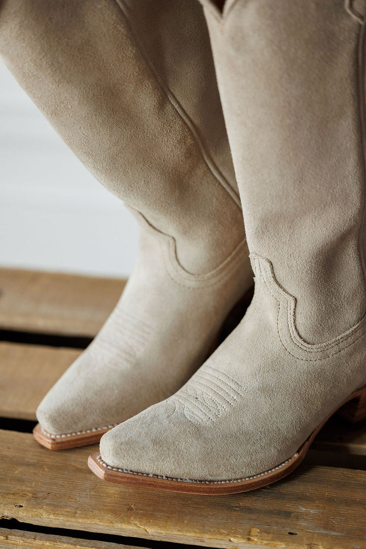 A close-up of The Shiloh western boots by Savannah Sevens western life{&}style, featuring light beige rough out leather, square toes, and decorative stitching, resting on a wooden surface. Made by Black Star. 