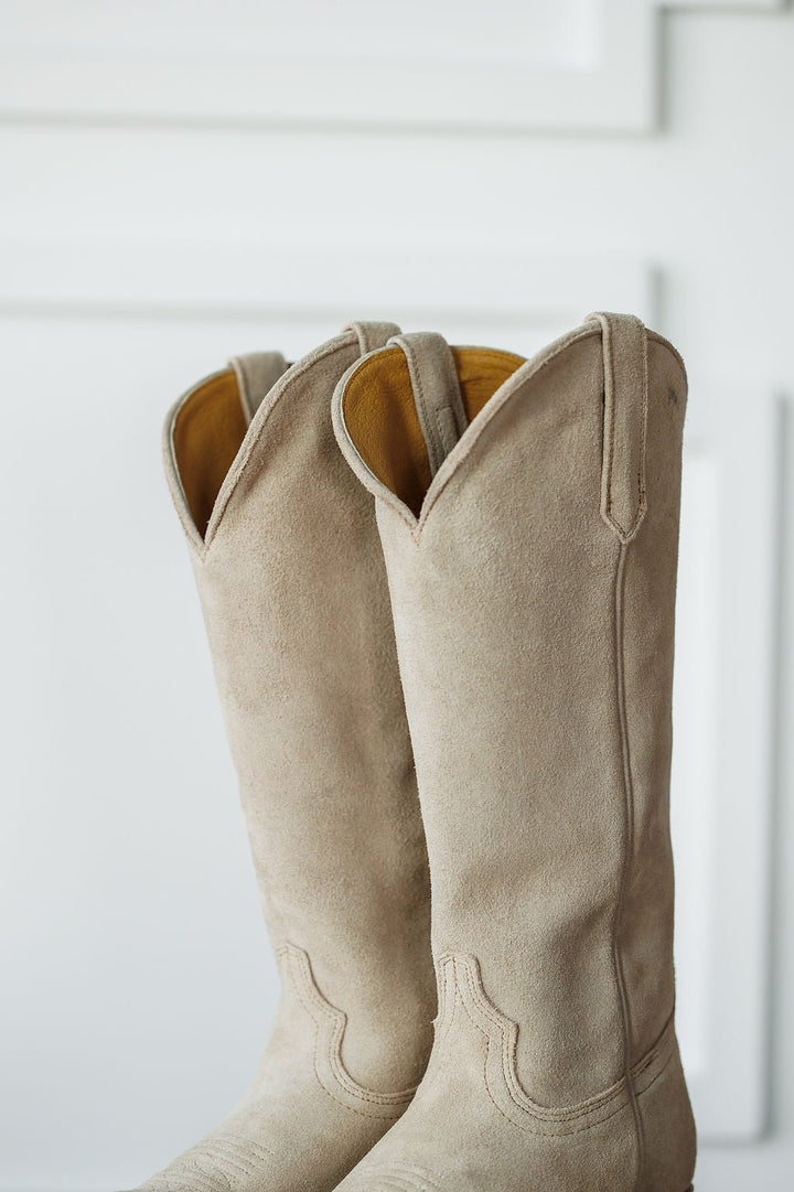 A close-up of The Shiloh boots by Savannah Sevens western life{&}style—tall, light beige rough out leather western boots with decorative stitching, snip toe, and curved tops—standing upright against a white background. Made by Black Star. 