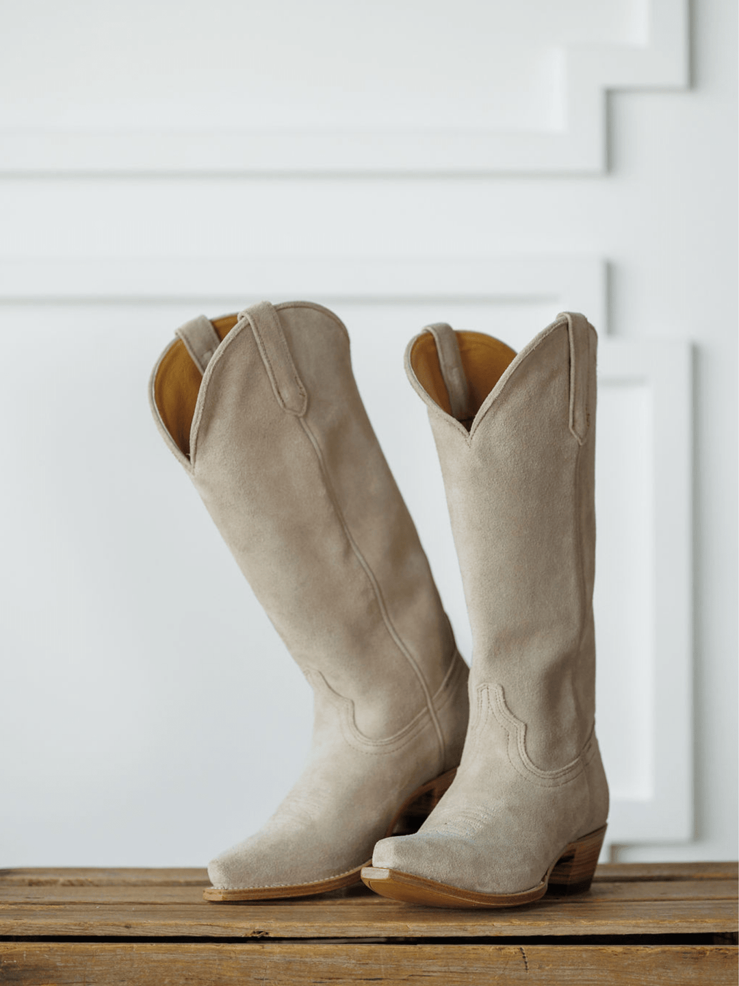 The Shiloh tall snip toe western boots by Savannah Sevens western life{&}style, in light beige with wooden heels, stand on a wooden surface against a white paneled background. Made by Black Star. 