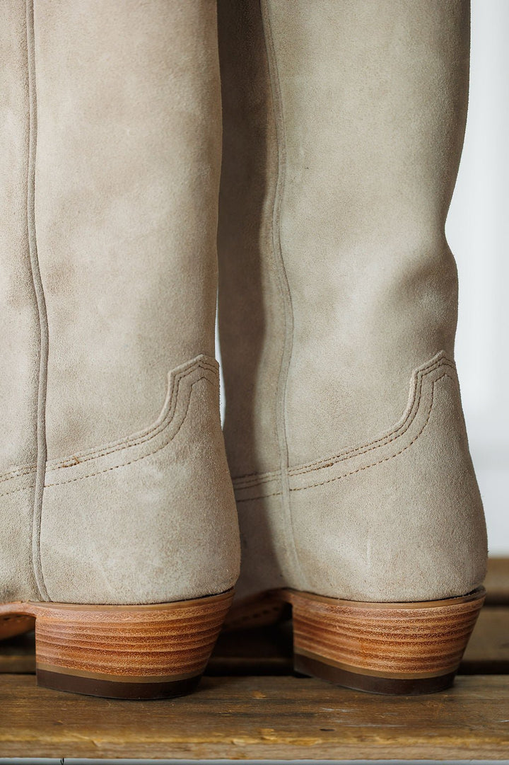 Close-up of the backs of two The Shiloh boots by Savannah Sevens western life{&}style in light beige rough out leather with wooden stacked heels, detailed ankle stitching, and a rustic wooden surface underneath. Made by Black Star. 