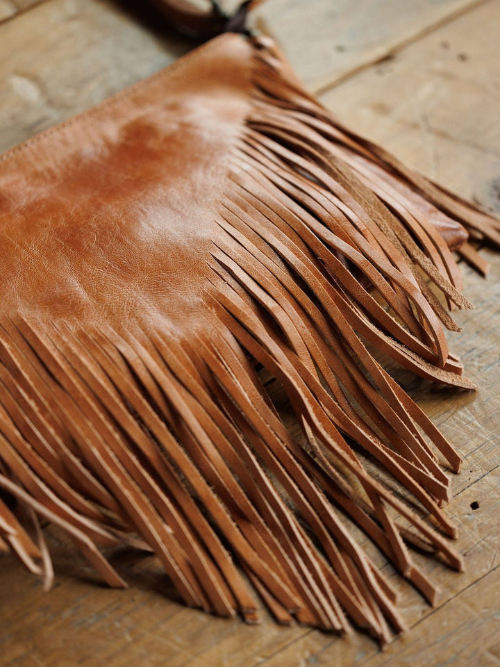 A close-up of The Tahama by Savannah Sevens western life{&}style, a fringe leather crossbody bag, rests on rustic wood with detailed long, thin fringe hanging from the bottom.
