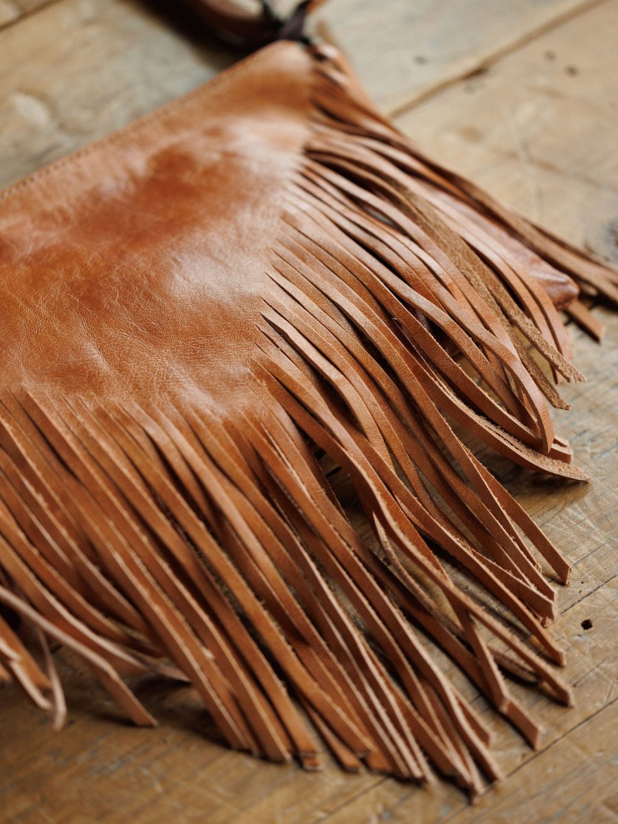 A close-up of The Tahama by Savannah Sevens western life{&}style, a fringe leather crossbody bag, rests on rustic wood with detailed long, thin fringe hanging from the bottom.