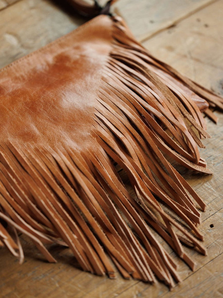 A close-up of The Tahama by Savannah Sevens western life{&}style, a fringe leather crossbody bag, rests on rustic wood with detailed long, thin fringe hanging from the bottom.