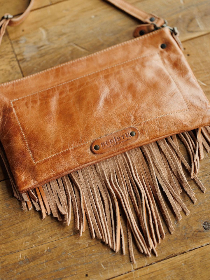 The Tahama by Savannah Sevens western life{&}style is a rectangular leather crossbody bag with fringe, visible stitching, and a metal nameplate shown on a rustic wooden surface.