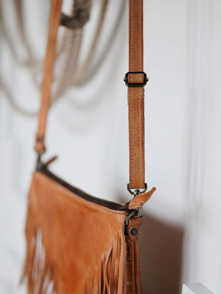 Close-up of The Tahama crossbody bag by Savannah Sevens western life{&}style, featuring intricate leather fringe and an adjustable strap with a metal buckle, hanging on a wall against a softly blurred background.