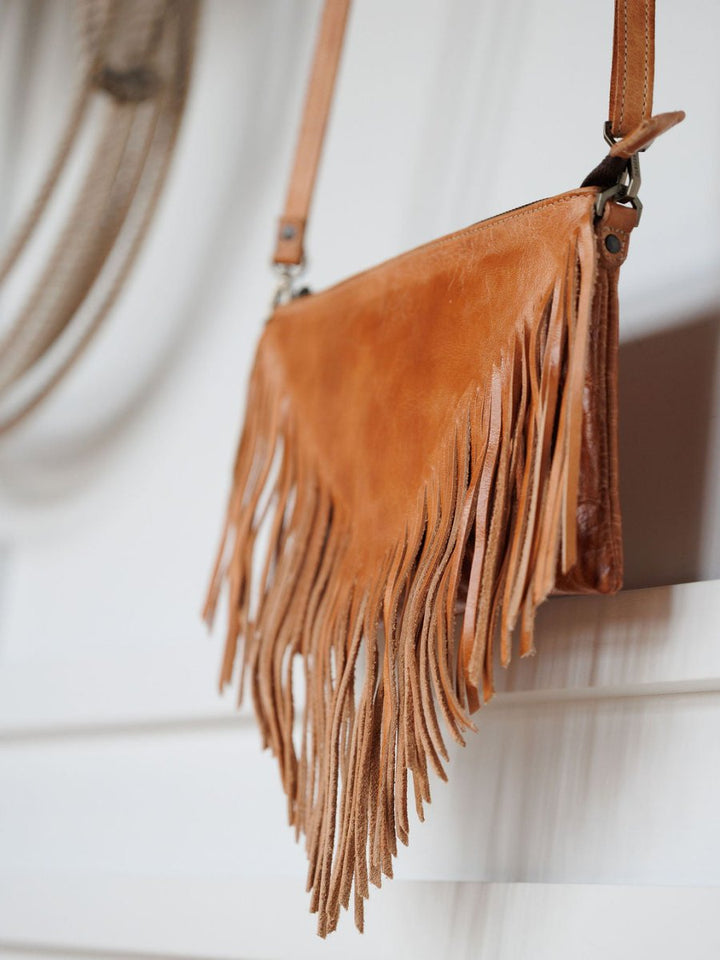 The Tahama by Savannah Sevens western life{&}style, a fringe leather crossbody bag with long tan fringe, hangs against a white wall for a bohemian, stylish vibe.