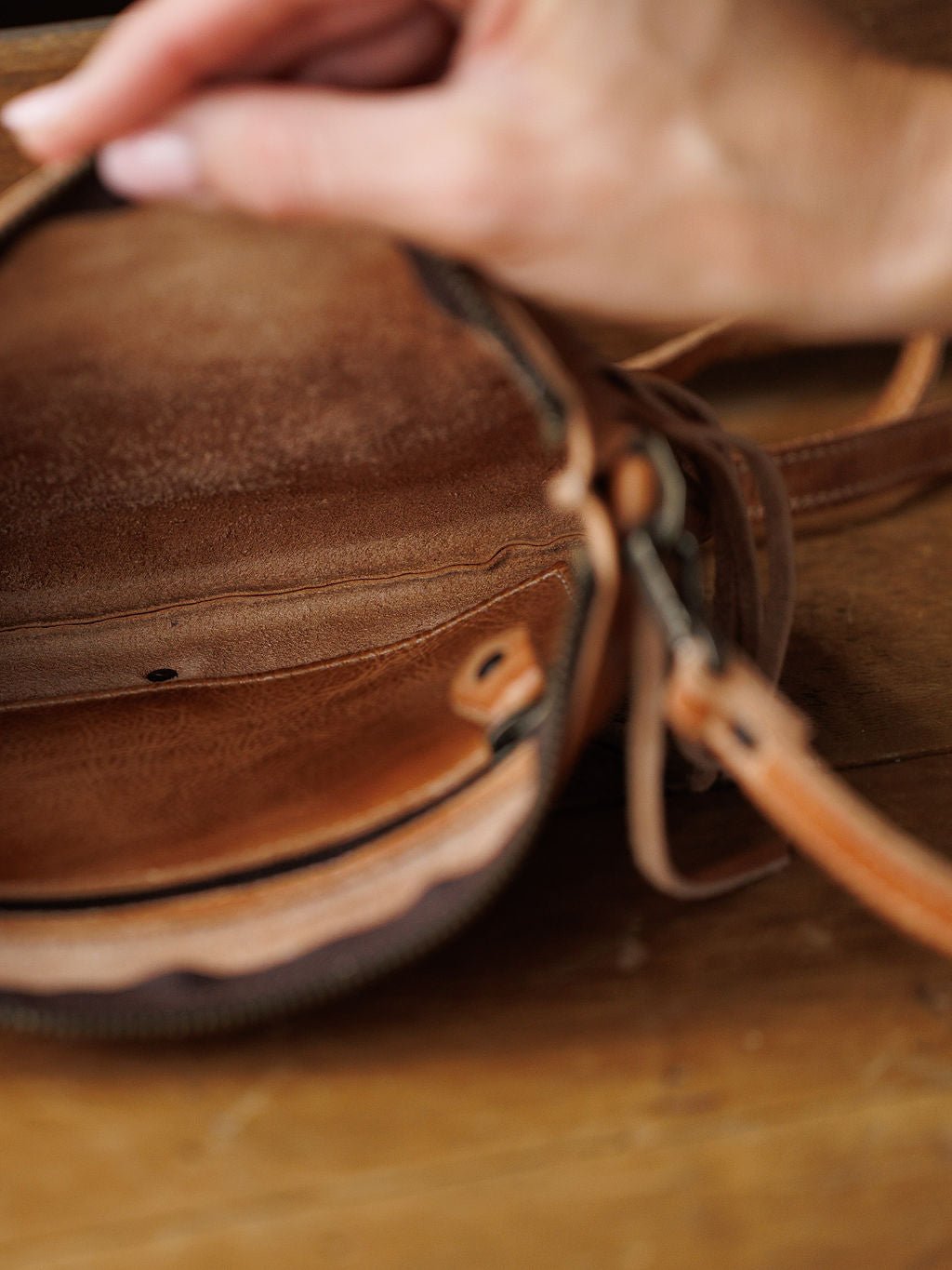A hand opens The Tahama clutch by Savannah Sevens western life{&}style, showing its empty, textured brown leather interior on a wooden surface.