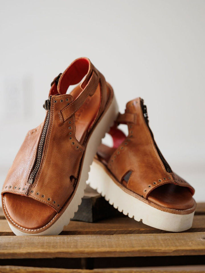 The Tayten by Savannah Sevens western life{&}style are distressed leather sandals with front zippers and white, thick, ridged soles, displayed on a wooden surface against a plain background.